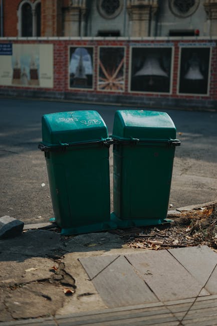 Two green waste bins with hinged lids are positioned side by side on the pavement at the edge of a street, with a small amount of leaf and soil debris collected at their bases. The bins are made of durable plastic with a textured surface, and are marked with white text that indicates their purpose for rubbish disposal. Behind the bins, there is a brick wall with partially visible storefronts that feature windows and signage, suggesting an urban environment. The ground surrounding the bins consists of concrete paving stones, some of which are slightly uneven, and a few stray twigs and leaves are scattered nearby. The street surface in front appears asphalt, with a curb separating it from the pavement. The overall scene reflects an area where private waste collection services, like those offered by House Clearance Lewisham, might be employed for rubbish removal, illustrating typical outdoor waste storage close to commercial or residential properties in Lewisham, London.
