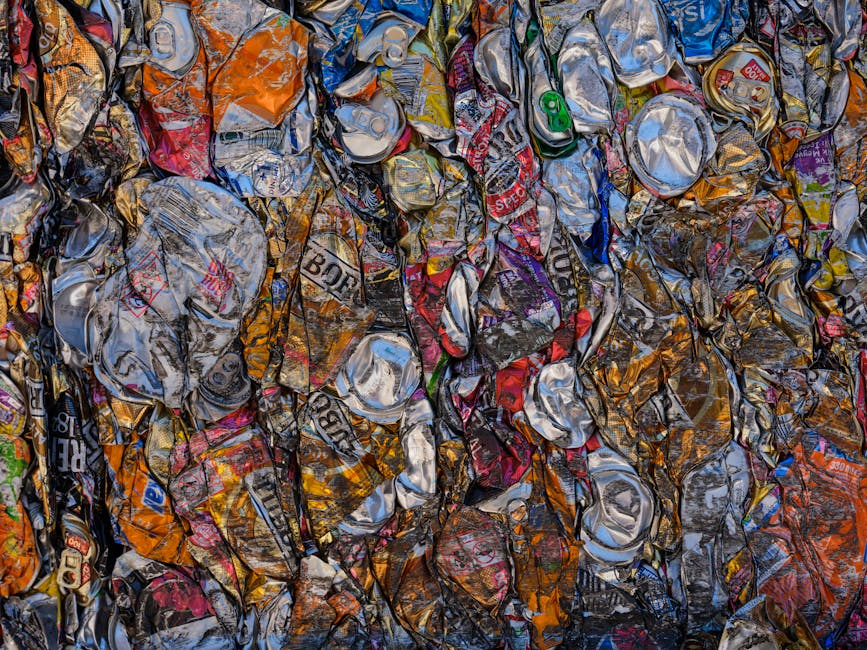 A densely packed pile of crushed aluminium cans and aluminium foil, resting on a flat surface. The cans display a variety of colours, including silver, red, blue, green, and orange, with some featuring brand labels and logos. The surface of the cans appears crumpled and torn, exhibiting a mix of shiny metallic textures and matte finishes, indicative of aluminium recycling. The cans are stacked irregularly, with some leaning against each other, and occupy the foreground of the image. In the background, a few other crushed beverage containers and fragments of aluminium waste can be seen, blending into the overall collection. The environment seems to be an indoor waste collection or recycling area, with no visible equipment or other contextual elements present. The lighting highlights the metallic sheen of the crushes, emphasizing their textured surfaces and the chaotic arrangement typical of bulk rubbish awaiting disposal or reuse, aligning with the context of independent waste handling and rubbish removal services offered by House Clearance Lewisham.