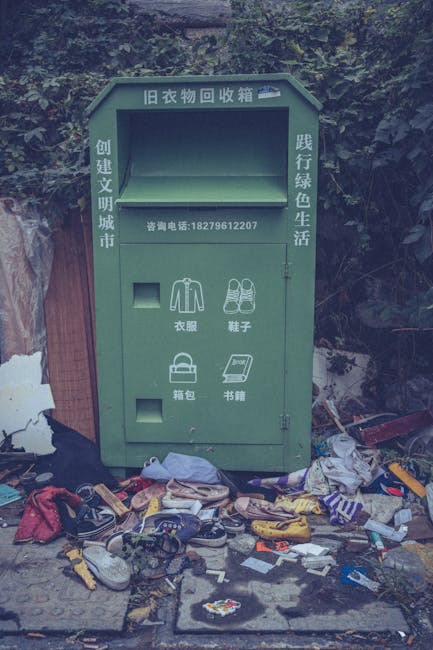 A green outdoor communal clothing donation and recycling bin positioned on a paved sidewalk, with a background of dense foliage and a wooden fence to the left. The bin features multiple compartments for different types of items, labeled in Chinese, including sections for clothing, shoes, bags, and books. It has a rectangular shape with a sloped top, and the label at the top indicates it is for recycling old clothes. In front of the bin, a large assortment of discarded items, primarily clothing, shoes, and miscellaneous trash, is spread across the ground; some garments are crumpled or scattered, with various colors and textures visible including textiles, plastic bags, and packaging. The scene appears untidy, suggesting a collection point for clothing reuse or donation that has been overwhelmed, emphasizing the importance of proper waste management or alternative disposal methods. This visual context relates to independent rubbish handling services, such as those offered by House Clearance Lewisham, in contrast with municipal waste collection, by highlighting the need for organized waste removal solutions in community settings.