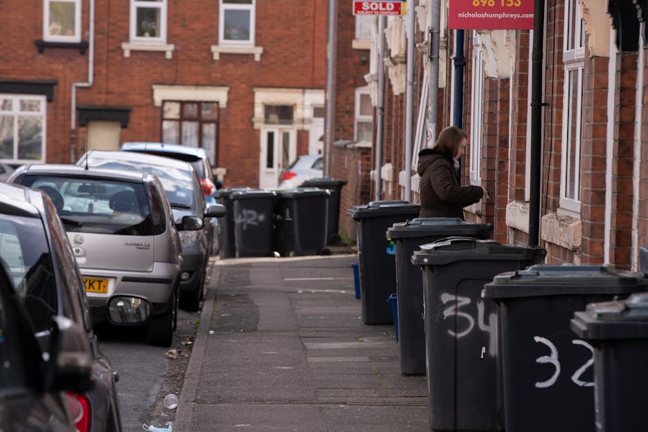 A residential street scene with a row of black wheelie bins placed along the sidewalk in front of a brick building. The bins are positioned close together, with some lined up against the building's exterior wall, which features multiple windows with white frames and stone sills. A woman dressed in a dark brown coat stands next to one of the bins, appearing to be opening or closing the lid. To the left, several parked cars, including a silver hatchback with a yellow license plate, are lined up along the curb, occupying part of the road and beyond. The environment appears to be in an urban area with terraced brick houses and a clean, paved sidewalk. The scene suggests on-site or private waste collection, which is an alternative to local authority rubbish disposal, as indicated by the location on a typical residential street, with the bins intentionally placed for collection. Natural daylight illuminates the scene, highlighting the textures of the brickwork, the plastic surfaces of the bins, and reflections on the car windows, contributing to a neutral and observational tone suitable for a rubbish removal service context.