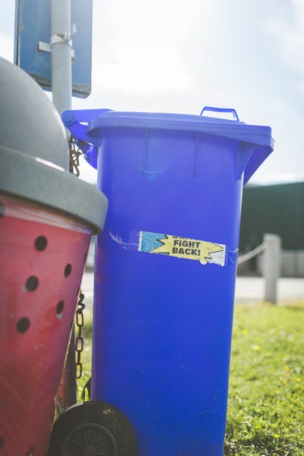 A bright blue wheeled rubbish bin with a closed lid, positioned outdoors on a patch of grass, features a small sticker with the words 'FIGHT BACK!' in yellow and blue. To its left, part of a larger red and pink polystyrene bin with black circular holes in the lid is visible, and a metal chain hangs from its side. In the background, a utility pole, a road barrier, and a cloudy sky are seen, suggesting a street or roadside environment. The scene appears to be part of a waste collection or rubbish disposal setting, with natural daylight illuminating the scene and highlighting the textures of the plastic bins and surrounding environment. This visual context aligns with private waste handling or rubbish removal services, such as those offered by House Clearance Lewisham, capable of on-site collection and alternative waste disposal solutions.