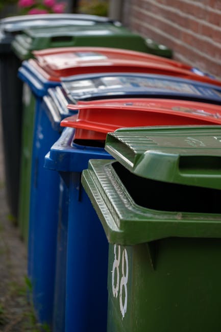 A residential street scene with a row of black wheelie bins placed along the sidewalk in front of a brick building. The bins are positioned close together, with some lined up against the building's exterior wall, which features multiple windows with white frames and stone sills. A woman dressed in a dark brown coat stands next to one of the bins, appearing to be opening or closing the lid. To the left, several parked cars, including a silver hatchback with a yellow license plate, are lined up along the curb, occupying part of the road and beyond. The environment appears to be in an urban area with terraced brick houses and a clean, paved sidewalk. The scene suggests on-site or private waste collection, which is an alternative to local authority rubbish disposal, as indicated by the location on a typical residential street, with the bins intentionally placed for collection. Natural daylight illuminates the scene, highlighting the textures of the brickwork, the plastic surfaces of the bins, and reflections on the car windows, contributing to a neutral and observational tone suitable for a rubbish removal service context.