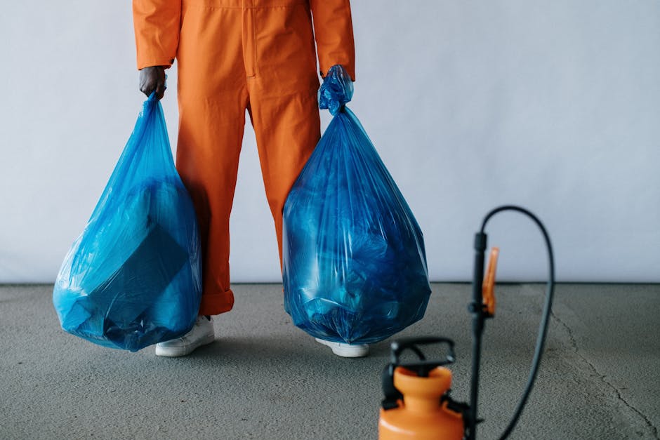 A person dressed in an orange work jumpsuit stands on a concrete floor, holding two large blue plastic rubbish bags filled with waste, one in each hand. The bags are semi-transparent, revealing crumpled and flattened recyclable materials inside. The individual's white footwear is visible beneath the pant cuffs. In the foreground, a pressure sprayer or cleaning device with an orange tank and black hose is partially visible, placed on the ground. The background consists of a plain, light-colored wall, providing a neutral setting typical of an indoor or covered area used for waste collection or disposal activities. The scene emphasizes the process of waste handling, consistent with the context of private rubbish removal or on-site clearance services. House Clearance Lewisham may arrange such waste collection, supporting alternative disposal methods outside local authority services, especially in commercial or residential settings requiring bulk rubbish removal.