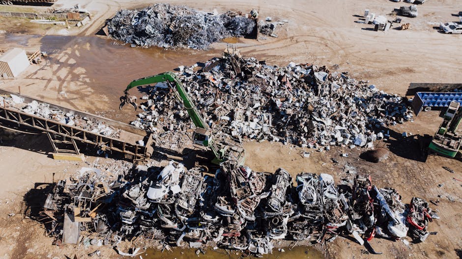 An aerial view of a large open area at a scrapyard or waste disposal site showing a pile of assorted scrap metal and debris, including twisted steel, broken appliances, and discarded vehicle parts. A green excavator with a mechanical claw is actively lifting and moving smaller sections of metal from the heap, positioned centrally within the scene. To the left, a long metal conveyor or ramp extends from the ground up to the scrap pile, assisting with the sorting process. Surrounding the main debris area, the ground is mostly bare, dusty, and uneven, with some patches of dirt and scattered smaller metal fragments. In the background, the area appears to be part of an industrial site with a few parked vehicles and additional piles of scrap material, all under bright natural daylight. The scene is centred on waste management activities related to recycling or destruction of metal objects, relevant to independent waste handling and rubbish removal services like those offered by House Clearance Lewisham.