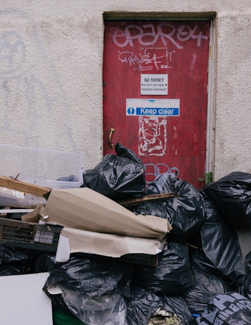 A pile of black garbage bags and discarded materials including cardboard and broken furniture are stacked against a textured, off-white exterior wall beneath a red metal door covered in graffiti and stickers. The door has various signs attached, one of which reads 'No Money' with additional small print, and another that says 'Keep Clear' in blue and white. The garbage bags and debris are on a pavement or outdoor area, indicating an uncollected rubbish accumulation likely associated with independent waste disposal or a private rubbish collection service. The overall scene suggests an area awaiting rubbish clearance or disposal, with environmental context typical of an urban setting where rubbish removal services like House Clearance Lewisham may be involved in managing such waste.