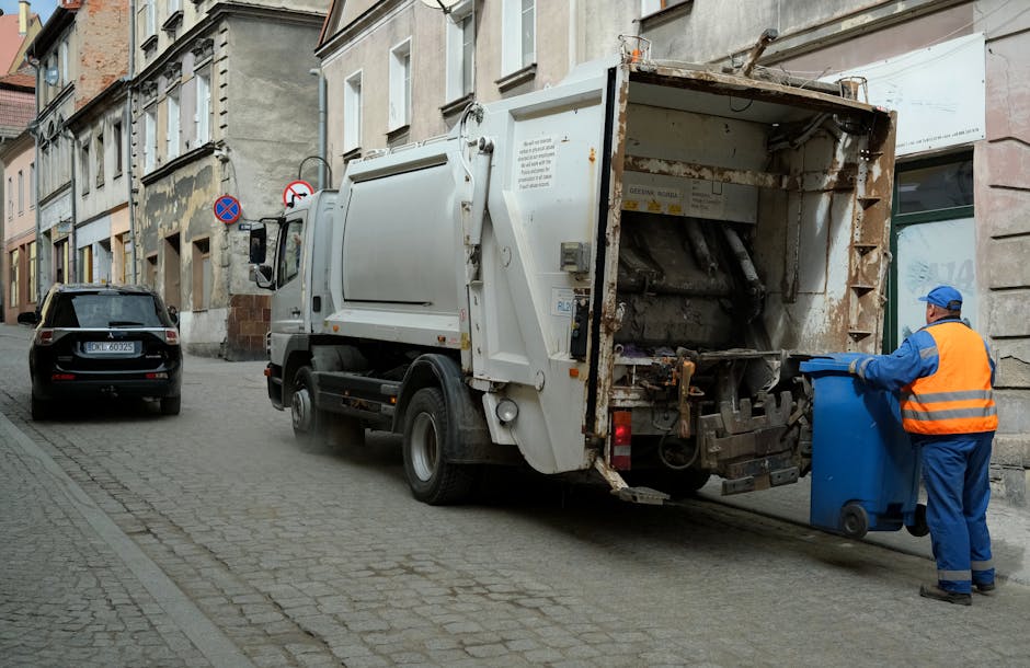 A pile of black garbage bags and discarded materials including cardboard and broken furniture are stacked against a textured, off-white exterior wall beneath a red metal door covered in graffiti and stickers. The door has various signs attached, one of which reads 'No Money' with additional small print, and another that says 'Keep Clear' in blue and white. The garbage bags and debris are on a pavement or outdoor area, indicating an uncollected rubbish accumulation likely associated with independent waste disposal or a private rubbish collection service. The overall scene suggests an area awaiting rubbish clearance or disposal, with environmental context typical of an urban setting where rubbish removal services like House Clearance Lewisham may be involved in managing such waste.