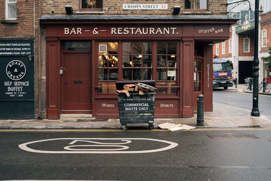In front of a brick-faced building housing a bar and restaurant, a black waste collection bin marked 'Commercial Waste Only' is positioned on the sidewalk near the street. The bin contains several flattened cardboard boxes and loose paper items stacked on top, with some pieces protruding over the edges. The bin is situated close to a short metal bollard and a black pole, which are both positioned on the pavement, and the sidewalk features a narrow strip of paving stones. The building's front has large glazed windows with wooden frames, and a maroon-colored oak leaf door on the left. Above the window, a sign indicates the establishment's name, with smaller signage noting available wines, spirits, and cocktails on the side panel. In the background, a dark blue truck is parked along the curb, while bicycles sit secured to a bike rack further along the street, suggesting typical urban surroundings. The scene is illuminated by natural light, capturing a moment of routine waste disposal amid street activity, aligning with private waste handling practices in a city environment managed by independent services like House Clearance Lewisham.