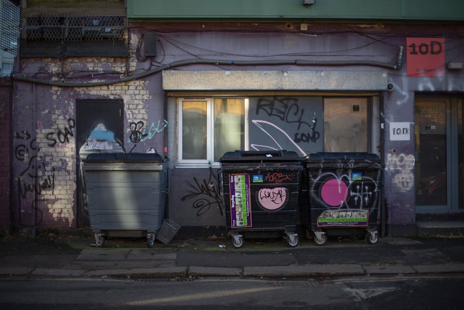 The image displays a row of three large wheelie bins positioned in front of a building with a brick and concrete exterior. The left bin is grey, slightly open at the top, and has a smooth, matte surface. The middle and right bins are black and feature various colourful stickers, graffiti tags, and symbols, including a large pink heart on each. The black bins are made of plastic with a textured surface and have hinged lids, with one lid partially open on the left black bin. They are situated on a concrete sidewalk next to a curb, with small patches of fallen leaves and debris scattered around. Behind the bins, a building wall shows signs of wear with patches of exposed brick, graffiti, and peeling paint. A small, closed window with frosted glass and a white frame is visible directly behind the bins, with graffiti tags and spray paint marked around it. To the right, there is an entrance door with a glass panel, also adorned with graffiti and stickers. The scene suggests urban surroundings where private waste collection services, such as those offered by House Clearance Lewisham, might be used for rubbish removal or house clearances, especially given the presence of graffiti and typical external waste storage arrangements.