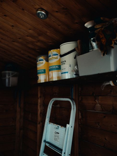 The image depicts a storage area with a dark wooden ceiling and wall panels. A horizontal shelf is mounted on the wall, holding several large cylindrical containers of sealing and protective products, primarily white with yellow and blue labels. One of the containers is partially visible, displaying the brand name 'Croseal.' To the right of these containers, a white rectangular box or container is placed, possibly for storing small tools or supplies, with some fabric or cloth draped over its edge. In the foreground, a small white step ladder is positioned against the wooden wall, partially extending into the frame. The lighting is dim, with a small recessed light fixture visible in the ceiling, casting soft illumination over the scene. The environment suggests a shed or storage room often used for DIY or maintenance items, fitting into the context of home improvement or waste management when clearing and disposing of unwanted materials like empty sealant containers and related supplies, which House Clearance Lewisham might handle through private disposal or rubbish collection services.