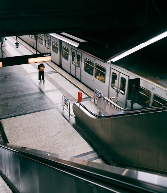 A view of an underground train station platform showing a silver commuter train with multiple windows, positioned on the right side of the image. The train's exterior is smooth and metallic with a slight reflective finish, and it is partially visible as it stops at the platform. On the left side, a solitary passenger wearing dark clothing and carrying a backpack is standing near a digital display board that shows station information, including the name 'Gleis 1'. The platform surface is made of light grey tiles with darker grey tactile paving strips near the edge. In the foreground, a stainless steel curved handrail runs along the escalator leading down to the platform area. Overhead lighting illuminates the scene evenly, creating a clean and modern ambiance typical of contemporary underground stations. An uplifted black sign above the platform indicates the station name and track number, subtly signalling the area's purpose for public transportation and the potential need for waste management services in station maintenance or cleanliness tasks. The environment appears tidy and well-maintained, with no visible litter or clutter, reflecting typical operational standards of urban rail infrastructure.