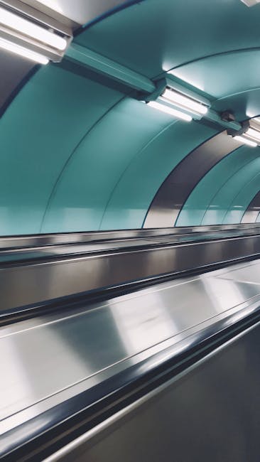 The image depicts a section of an underground or indoor transportation station with a modern, curved teal-colored ceiling and smooth metallic surfaces. In the foreground, there are two parallel, polished stainless steel handrails or barriers, suggesting the presence of an escalator or moving walkway. Fluorescent lights embedded in the ceiling illuminate the area evenly, highlighting the sleek finishes and clean environment. The teal paneling on the ceiling creates a contemporary aesthetic, with the metallic elements providing a reflective quality. The setting is free of clutter, and the environment appears well-maintained. This scene exemplifies a typical urban transit environment, where private or independent waste collection services like House Clearance Lewisham could be relevant for maintaining cleanliness and managing rubbish removal in transportation hubs or related infrastructure, especially during station renovations or upgrades. The overall atmosphere is neutral, professional, and organized, emphasizing the importance of cleanliness and efficient waste handling in public transport contexts.