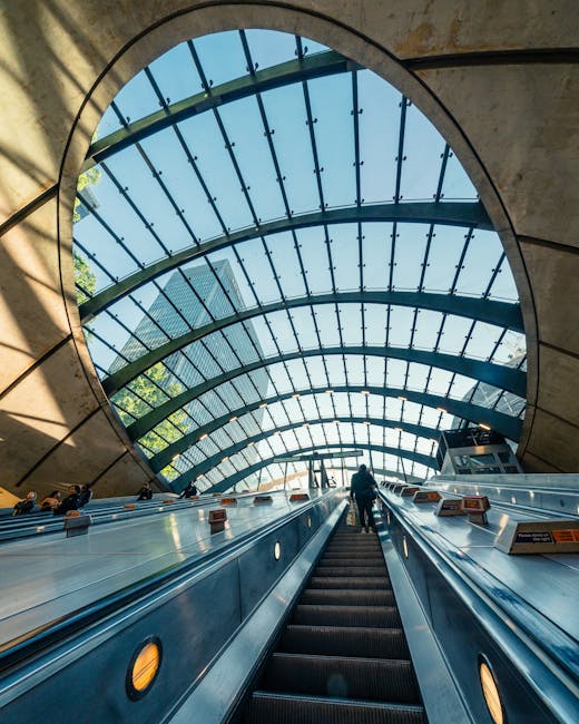 A view of an underground train station platform showing a silver commuter train with multiple windows, positioned on the right side of the image. The train's exterior is smooth and metallic with a slight reflective finish, and it is partially visible as it stops at the platform. On the left side, a solitary passenger wearing dark clothing and carrying a backpack is standing near a digital display board that shows station information, including the name 'Gleis 1'. The platform surface is made of light grey tiles with darker grey tactile paving strips near the edge. In the foreground, a stainless steel curved handrail runs along the escalator leading down to the platform area. Overhead lighting illuminates the scene evenly, creating a clean and modern ambiance typical of contemporary underground stations. An uplifted black sign above the platform indicates the station name and track number, subtly signalling the area's purpose for public transportation and the potential need for waste management services in station maintenance or cleanliness tasks. The environment appears tidy and well-maintained, with no visible litter or clutter, reflecting typical operational standards of urban rail infrastructure.