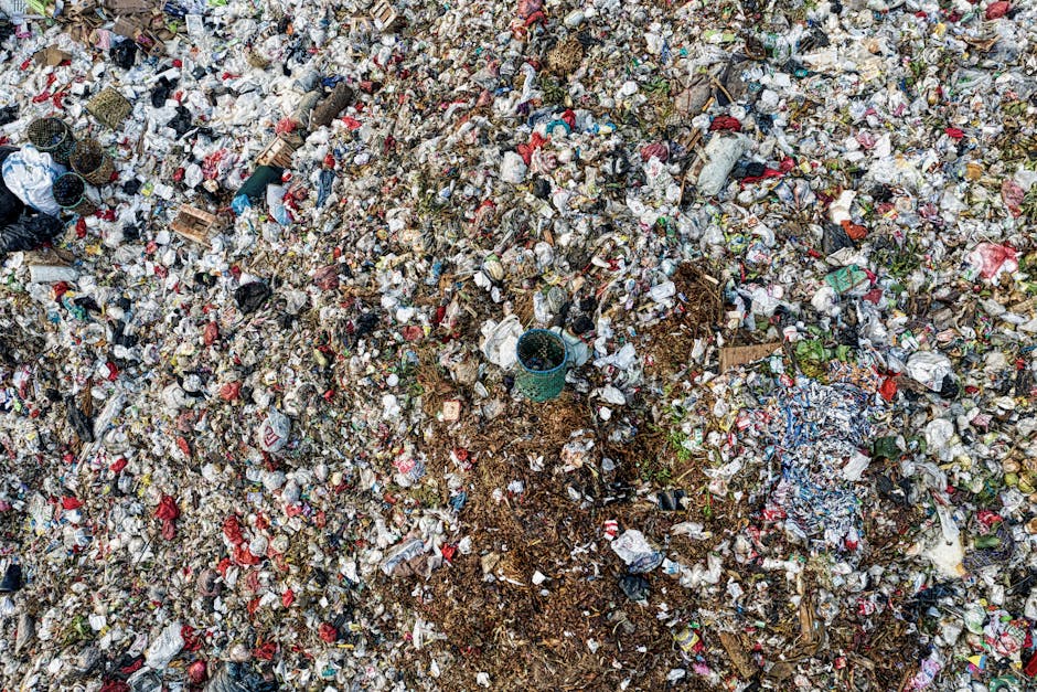 A high-angle view of a large landfill site filled with a dense layer of mixed waste materials. The refuse includes plastic bottles, food packaging, cardboard, paper, and other household waste, creating a patchwork of various colors and textures. In the center, a small green plastic bucket is partially buried among the rubbish, with nearby earth and soil visible where waste has been pushed aside or disturbed. Surrounding the waste, there are small piles of dirt and organic debris, contrasting with the synthetic, crumpled, and shredded plastics and paper. The surface appears uneven and compacted, with some items partially buried or overlapped, illustrating an unmanaged or natural landfill environment. The lighting is diffuse, with no strong shadows, emphasizing the chaotic and cluttered appearance typical of waste heaps. This scene reflects the context of rubbish accumulation, where independent waste handling or collection by specialists such as House Clearance Lewisham may be necessary for proper removal or site clearance, especially given the extent and disorder of the waste pile.