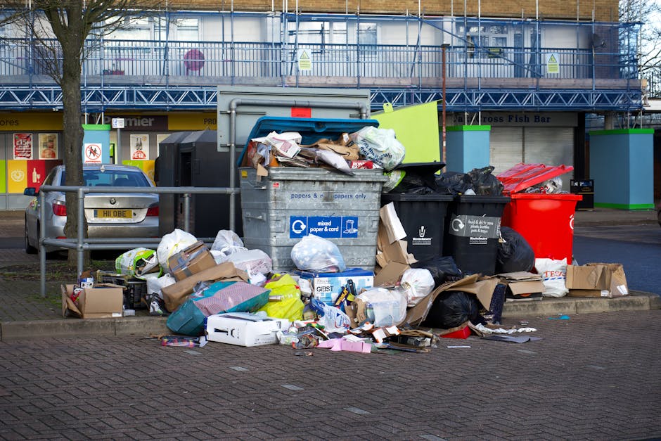 A collection of overflowing rubbish bins and numerous scattered waste items on a town street, including cardboard boxes, plastic bags, paper packaging, and miscellaneous household waste. The central grey bin appears to be designated for mixed paper and cardboard, with its lid open, overflowing with crumpled papers, cardboard pieces, and rejected packaging. To its right are black bins filled with plastic and other waste, along with a bright red bin nearby, also piled high with discarded rubbish. Several paper bags and open cardboard boxes are on the paved ground, with various crumpled papers, plastic bottles, and small packaging items strewn around. In the background, there is a row of small retail shops with closed shutters, and an off-white building with scaffolding and safety signs, indicating possible ongoing maintenance. A parked grey car is partially visible on the left side, and a metal railing separates the waste area from the parking spaces. The scene reflects a typical urban setup where waste has accumulated outside the designated bins, relating to private waste management or alternative collection services like those offered by House Clearance Lewisham.