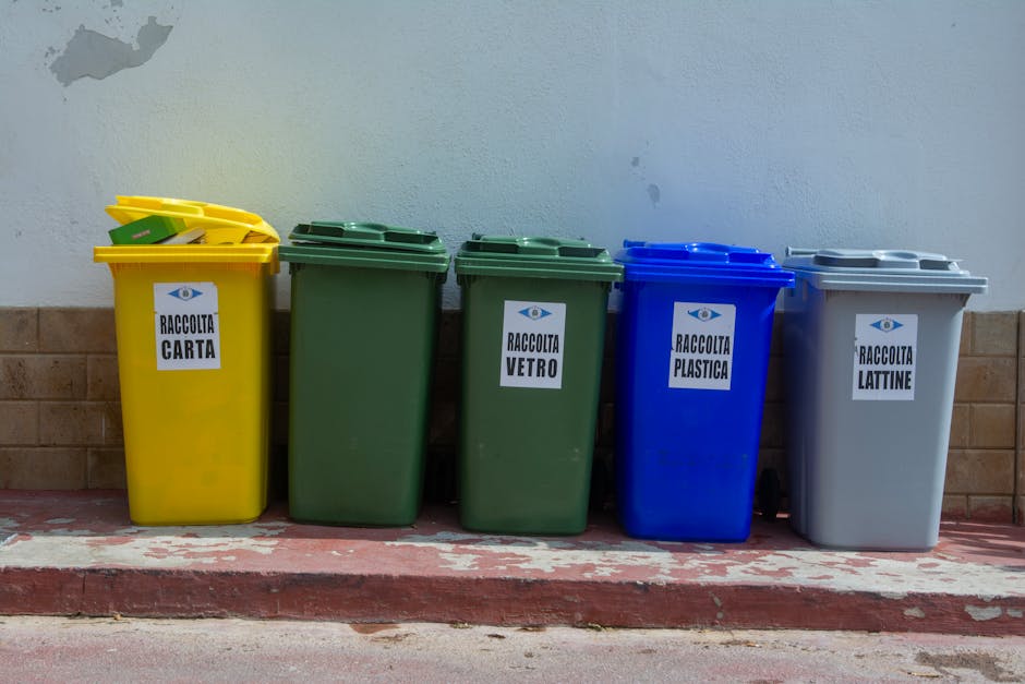 A high-angle view of a large landfill site filled with a dense layer of mixed waste materials. The refuse includes plastic bottles, food packaging, cardboard, paper, and other household waste, creating a patchwork of various colors and textures. In the center, a small green plastic bucket is partially buried among the rubbish, with nearby earth and soil visible where waste has been pushed aside or disturbed. Surrounding the waste, there are small piles of dirt and organic debris, contrasting with the synthetic, crumpled, and shredded plastics and paper. The surface appears uneven and compacted, with some items partially buried or overlapped, illustrating an unmanaged or natural landfill environment. The lighting is diffuse, with no strong shadows, emphasizing the chaotic and cluttered appearance typical of waste heaps. This scene reflects the context of rubbish accumulation, where independent waste handling or collection by specialists such as House Clearance Lewisham may be necessary for proper removal or site clearance, especially given the extent and disorder of the waste pile.