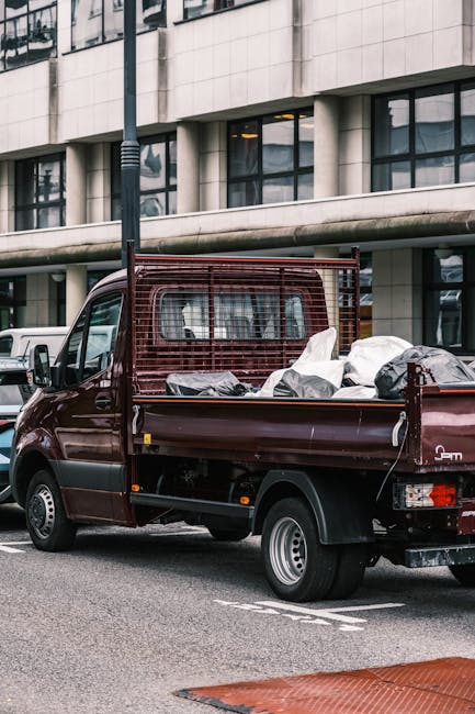 A compact burgundy pickup truck parked on a city street in front of a modern office building with large glass windows and beige stone cladding. The truck's bed is loaded with various rubbish bags and miscellaneous waste materials, some appearing to be black plastic sacks, while others are light-colored or transparent, revealing contents inside. The truck features a protective metal grid at the front of the cargo area, and the bed has metal sides with visible bolts. The rear wheel is visible, showing a silver rim and black tire. In the background, part of another vehicle can be seen along with a streetlight, and the overall scene suggests ongoing rubbish collection or clearance activity, reflecting the role of private waste handling services such as those offered by House Clearance Lewisham. The environment is urban, with the scene set during daylight hours, and the surrounding architecture indicating a commercial or office district, where independent rubbish removal is often required for property clearance or decluttering projects.