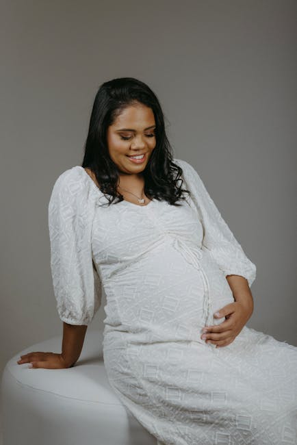 This image shows a pregnant woman with medium skin tone, dark shoulder-length hair, and a gentle smile, seated on a white surface against a plain neutral background. She is wearing a white, long-sleeved, textured dress that conforms to her baby bump, with subtle geometric patterns and a modest neckline. Her right hand is resting on the surface beside her while her left hand is lightly placed on her abdomen, highlighting her pregnancy. The lighting is soft and even, illuminating her face and dress without harsh shadows, creating a calm and composed atmosphere. The absence of environmental details suggests an indoor studio setting. Although no rubbish removal or waste context is visible in this specific image, the professional studio environment and the neutral background support visual consistency with content related to health and well-being, contrasting with the typical raw or cluttered scenes associated with rubbish collection or clearance activities, aligning more with promotional or personal documentation purposes.