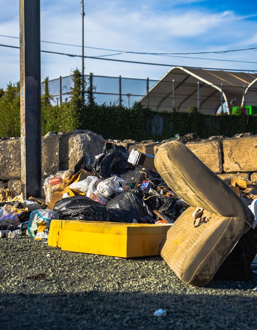 A pile of mixed household rubbish is situated on a gravel surface near a stone wall and a metal utility pole, featuring black and clear plastic garbage bags, a yellow plastic container, and an upturned, weathered car tire. Behind the rubbish, a rugged stone wall separates the area from a background of greenery, including trees and shrubs, with a partially visible tennis court enclosed by a dark wire mesh fence. The sky is clear with a few clouds, and overhead power lines run across the scene, indicating an outdoor urban or suburban environment. The scene reflects an instance of informal rubbish disposal, which may be managed through private waste collection services like those offered by House Clearance Lewisham, instead of standard municipal collection, highlighting the importance of proper waste removal methods for local property clearance or alternative rubbish handling. Overall, the image emphasizes the types of household waste that can accumulate outside designated collection points, illustrating potential access challenges for rubbish removal services in Lewisham.